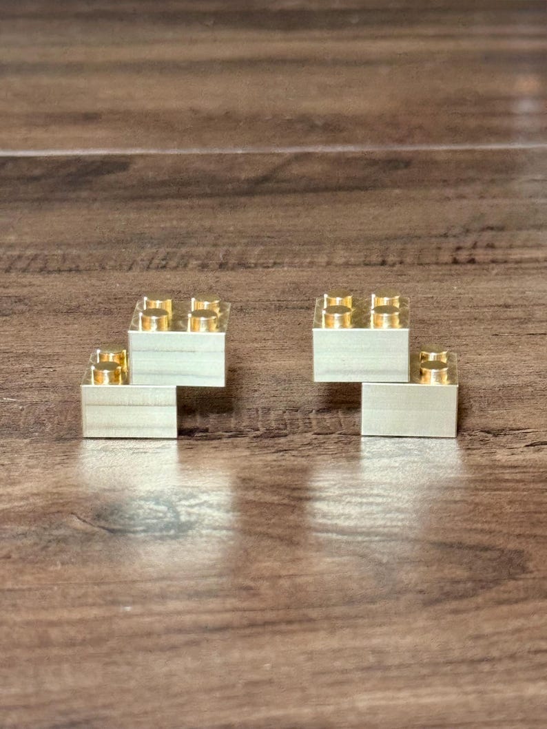 May include: Two metallic gold-colored building blocks, each featuring a rectangular base with four circular studs on the top surface. The blocks are arranged in a stepped configuration, showcasing a smooth, reflective finish and precise geometric edges.
