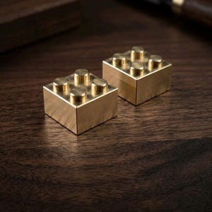 May include: Two square-shaped building blocks crafted from polished brass. Each block features four circular studs on the top surface, arranged in a two-by-two grid pattern. The metallic surfaces have a smooth, reflective finish with visible golden tones.