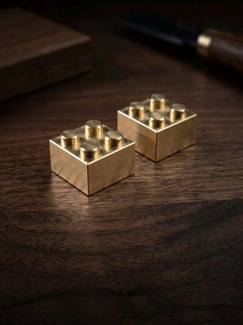 May include: Two square-shaped building blocks crafted from polished brass. Each block features four circular studs on the top surface, arranged in a two-by-two grid pattern. The metallic surfaces have a smooth, reflective finish with visible golden tones.