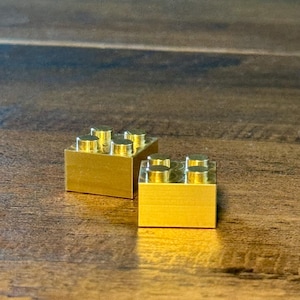 May include: Two rectangular building blocks made of polished gold-colored metal. Each block features four circular studs on the top surface, arranged in a two-by-two grid pattern, reflecting light with a smooth, metallic finish and sharp edges.