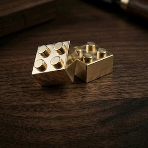 May include: Two metallic, gold-colored building blocks, each featuring a square shape with four raised circular studs on the top surface. The blocks have a smooth, polished finish and are positioned near each other, reflecting light on their surfaces.