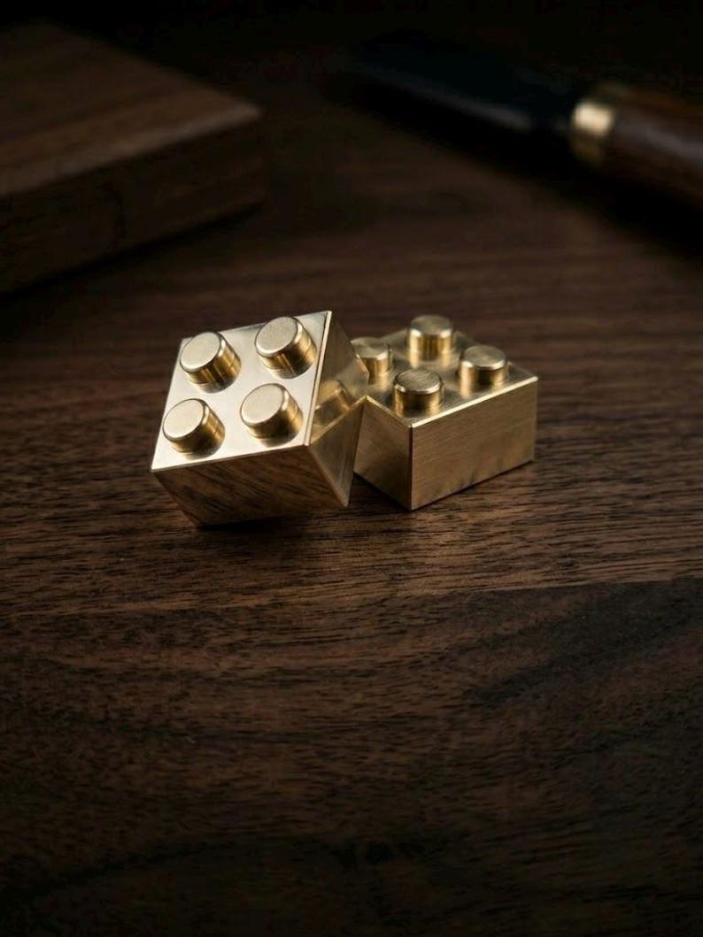 May include: Two metallic, gold-colored building blocks, each featuring a square shape with four raised circular studs on the top surface. The blocks have a smooth, polished finish and are positioned near each other, reflecting light on their surfaces.