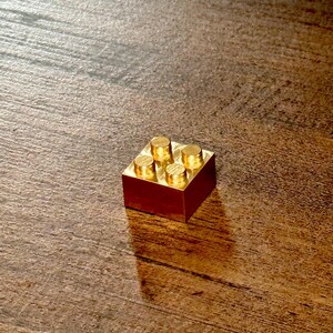 May include: A small, square, metallic gold-colored building block featuring four circular studs on its top surface. The object has a smooth, reflective finish and a solid, blocky construction, appearing as a single unit with a flat base.