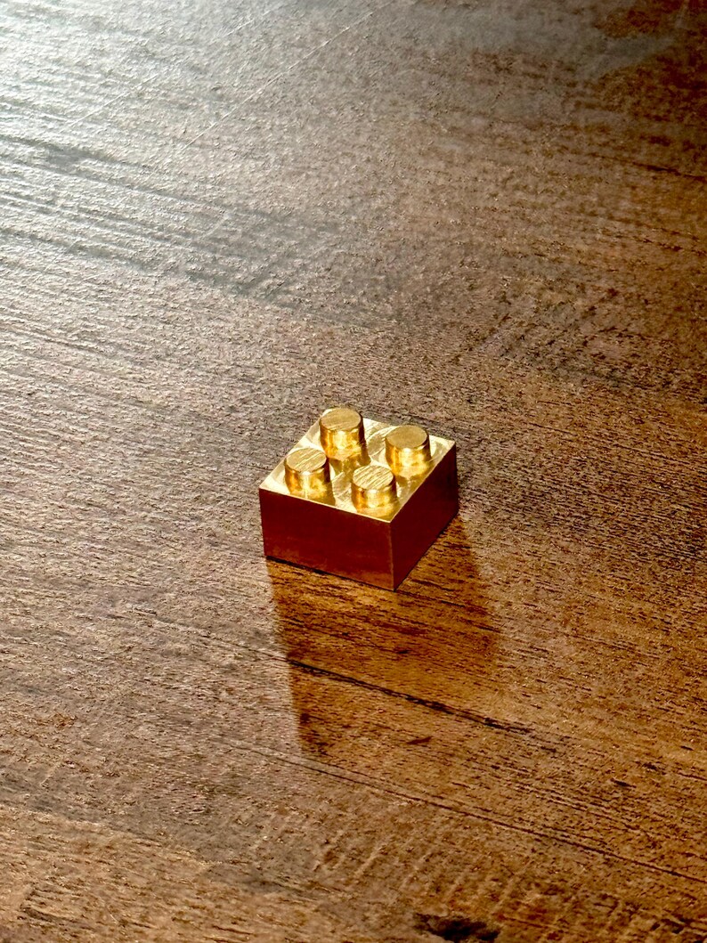 May include: A small, square, metallic gold-colored building block featuring four circular studs on its top surface. The object has a smooth, reflective finish and a solid, blocky construction, appearing as a single unit with a flat base.