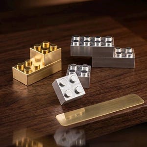 May include: A collection of metallic building blocks in gold and silver tones. The set includes rectangular and square blocks with circular studs on top, arranged in various configurations, resting on a dark wooden surface with a brass plate.