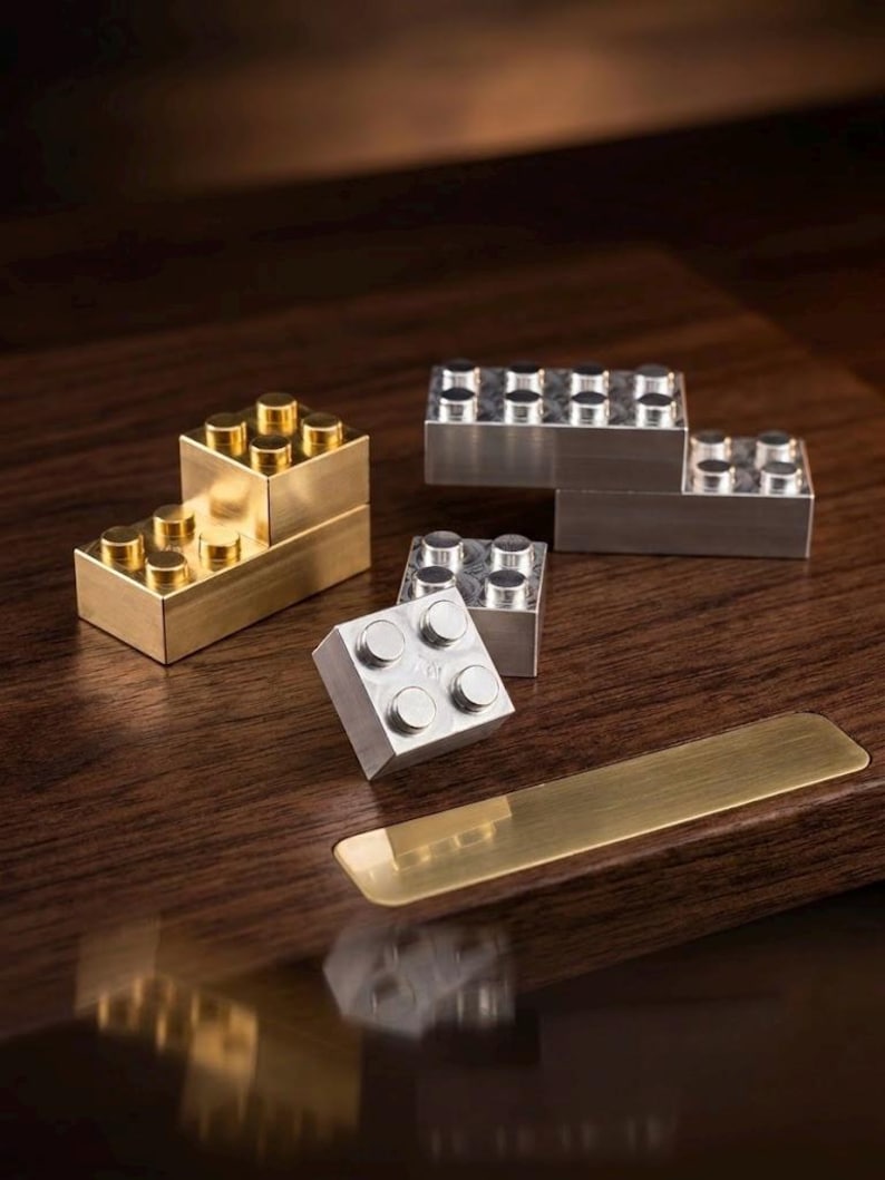 May include: A collection of metallic building blocks in gold and silver tones. The set includes rectangular and square blocks with circular studs on top, arranged in various configurations, resting on a dark wooden surface with a brass plate.