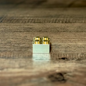 May include: A single rectangular building block made of polished gold-colored metal. The block features four circular studs arranged in a two-by-two grid on the top surface, reflecting light with a smooth, metallic finish and clean edges.