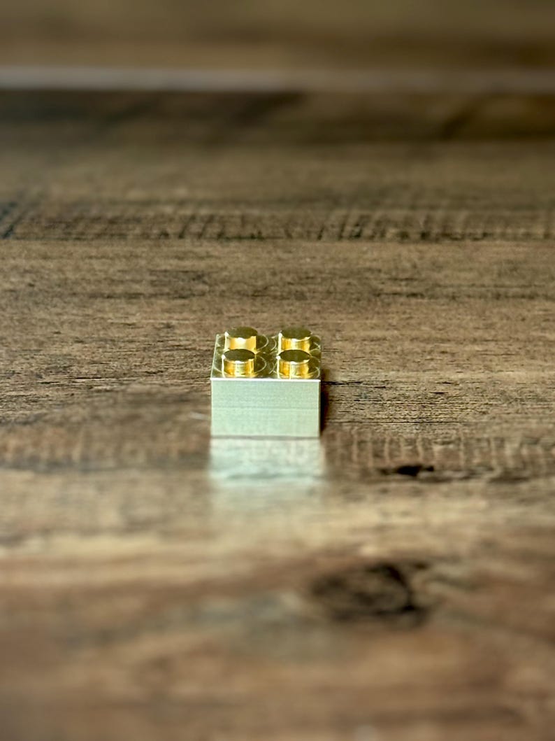 May include: A single rectangular building block made of polished gold-colored metal. The block features four circular studs arranged in a two-by-two grid on the top surface, reflecting light with a smooth, metallic finish and clean edges.