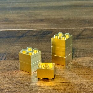 May include: Three metallic gold-colored building blocks made of solid material. One stack consists of two blocks, another stack consists of three blocks, and one single block is positioned in the front with its hollow underside facing upward.