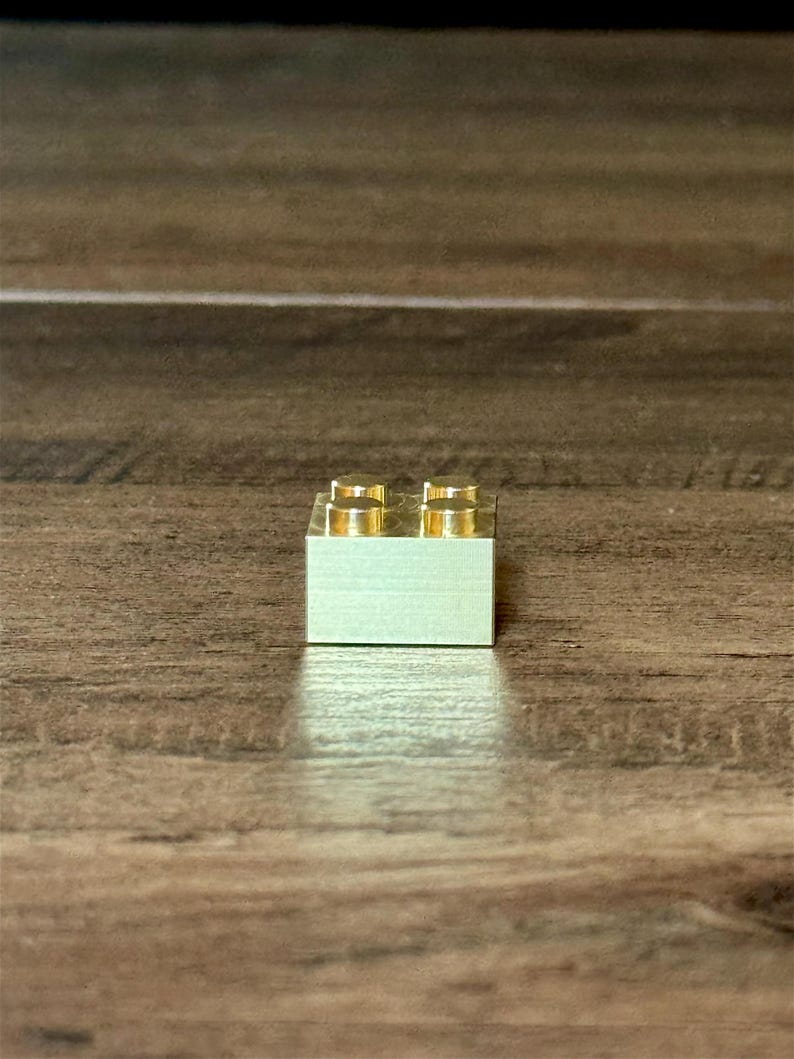 May include: A small, rectangular, metallic gold-colored building block featuring four circular studs arranged in a two-by-two grid on its top surface. The object has a smooth, reflective finish and a solid, blocky construction with sharp edges.