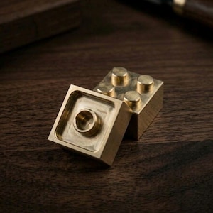 May include: Two interlocking building blocks made of polished brass. One block is positioned horizontally, displaying four circular studs on its top surface, while the other block is tilted, revealing a hollow square underside with a central circular connector.