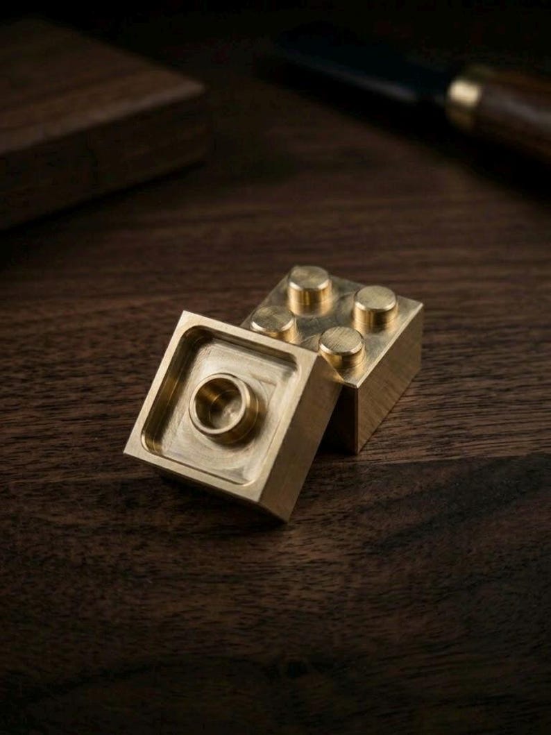 May include: Two interlocking building blocks made of polished brass. One block is positioned horizontally, displaying four circular studs on its top surface, while the other block is tilted, revealing a hollow square underside with a central circular connector.