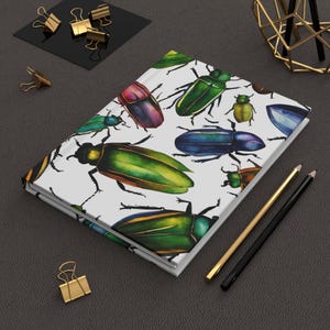 May include: A white notebook with a colorful beetle pattern. The beetles are green, blue, red, and yellow. The notebook is lying on a dark gray surface with two pencils and a gold metal stand.