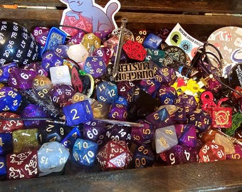 May include: A dark wooden chest filled with a large collection of multi-sided polyhedral dice in various colors, including purple, blue, and red. The chest contains a large black 100-sided die, a cat sticker, a sword charm, and a Dungeon Master sign.