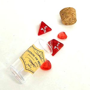 Peut inclure: Une fiole en verre transparent avec un bouchon en liège et une étiquette beige portant l'inscription POTION OF HEALING premium quality 2d4+2. Deux dés rouges translucides à quatre faces avec des chiffres blancs et deux petits cœurs en acrylique rouge.