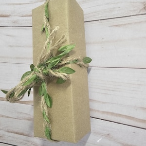 A rectangular gift box wrapped in thick kraft paper, adorned with a twine and green leaf ribbon. The box is positioned diagonally against a light wood-grain background, creating a rustic aesthetic. A beautifully wrapped gift is in thick Kraft paper, and tied with a hemp and green leaf ribbon.