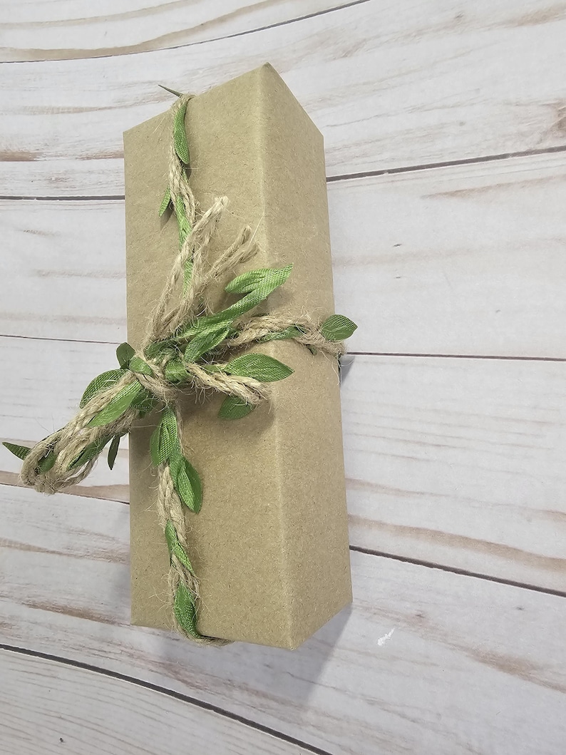 A rectangular gift box wrapped in thick kraft paper, adorned with a twine and green leaf ribbon. The box is positioned diagonally against a light wood-grain background, creating a rustic aesthetic. A beautifully wrapped gift is in thick Kraft paper, and tied with a hemp and green leaf ribbon.