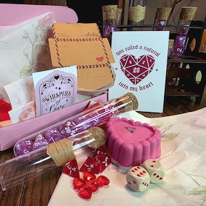 May include: A pink gift box containing a card with the text 'you rolled a natural 20 into my heart' and a 'Whispers of Love' tag. Includes glass vials with purple and red polyhedral dice, a pink heart-shaped wax item, and two white dice.