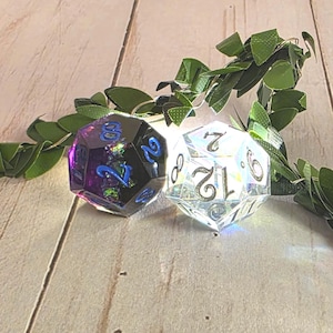 Two sharp-edge D12 polyhedral dice for Daggerheart tabletop gaming. One die is a deep obsidian black with swirling purple and blue nebula accents, and the other is crystal clear with high-contrast silver numbers. The dice are positioned on a wooden surface with vibrant green forest leaves in the background, highlighting the sensory prop-master aesthetic.