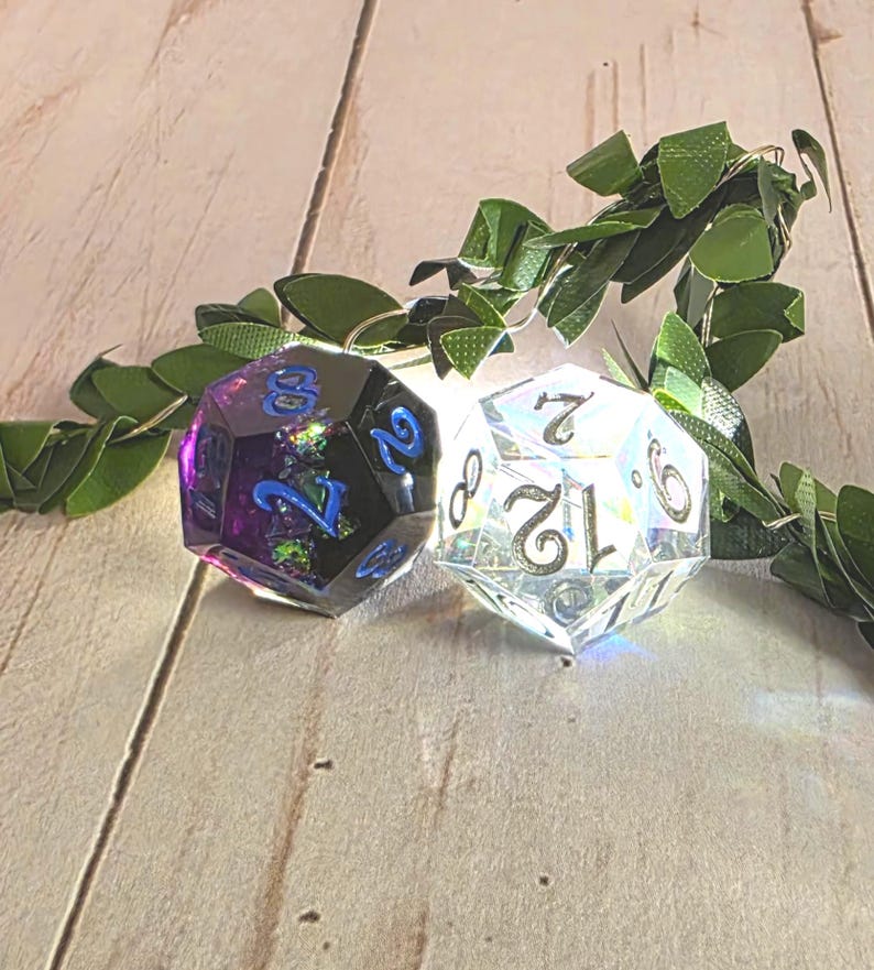 Two sharp-edge D12 polyhedral dice for Daggerheart tabletop gaming. One die is a deep obsidian black with swirling purple and blue nebula accents, and the other is crystal clear with high-contrast silver numbers. The dice are positioned on a wooden surface with vibrant green forest leaves in the background, highlighting the sensory prop-master aesthetic.