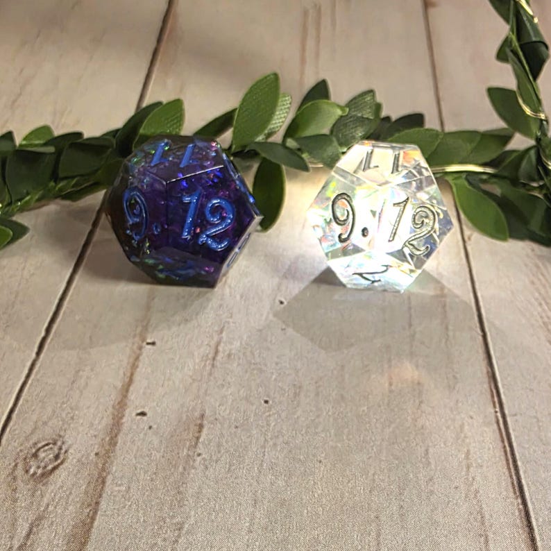 Puede incluir: Dos dados de doce caras. Uno es de color p&uacute;rpura oscuro con n&uacute;meros azules, el otro es transparente con brillo iridiscente y n&uacute;meros negros. Los dados est&aacute;n colocados sobre un fondo de hojas verdes y una superficie de madera clara.