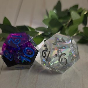 Puede incluir: Dos dados poli&eacute;dricos. Uno es de color p&uacute;rpura oscuro con n&uacute;meros azules, el otro es transparente con brillo iridiscente y n&uacute;meros negros. Los dados son para juegos de mesa.