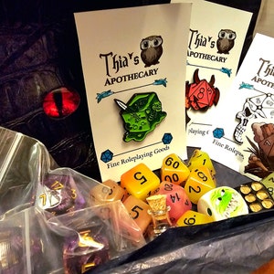 May include: A collection of colourful dice and a small glass bottle with a cork stopper. The dice are in a variety of colours, including yellow, orange, pink, green, and purple. There are also three pins featuring dice with different designs. The pins are on cards with the text "Thia's Apothecary Fine Roleplaying Goods".