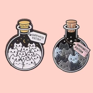Peut inclure: Deux épingles en émail en forme de flacons de potion, remplis de visages de chats. L'un est noir avec des chats blancs, l'autre noir avec des chats gris. Les deux ont un bouchon en liège et une étiquette "Happiness Potion".