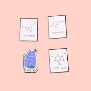 May include: Four enamel pins on a pink background. Three pins are rectangular with white backgrounds and black text and diagrams for Serotonin, Dopamine, and Caffeine. The fourth pin is a blue mole in a beaker with the number 6.022 x 10^23.