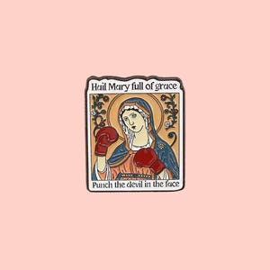 May include: A square enamel pin featuring an illustration of a religious figure wearing boxing gloves. The pin includes the text "Hail Mary full of grace" at the top and "Punch the devil in the face" at the bottom. The figure is surrounded by a gold halo.