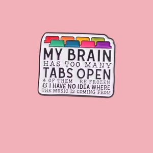May include: A white enamel pin with the text "MY BRAIN HAS TOO MANY TABS OPEN" and colorful tabs on top. The pin also includes the text "4 OF THEM RE FROZEN & I HAVE NO IDEA WHERE THE MUSIC IS COMING FROM".