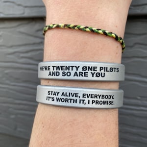 Puede incluir: Dos pulseras de silicona plateadas con texto negro. La pulsera superior dice "We're Twenty One Pilots and so are you." La pulsera inferior dice "Stay alive, everybody. It's worth it, I promise."