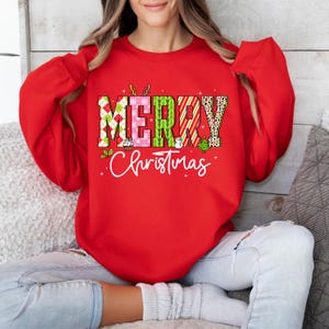 Può includere: Una felpa rossa con le parole "MERRY Christmas" in un design colorato e fantasia. La parola "MERRY" presenta un mix di motivi a quadri, rosa e leopardati. La parola "Christmas" è scritta in corsivo.