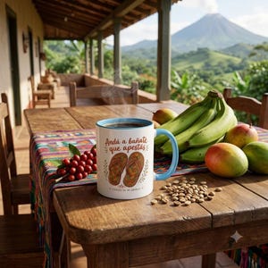 May include: A white ceramic mug with a light blue rim and handle, featuring a graphic of two woven sandals and the text "Andá a bañate que apestás" and "FRASES DE MI ABUELA" in brown, filled with dark coffee and emitting a thin wisp of steam.