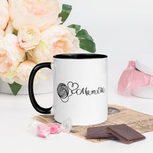 Op de afbeelding: Witte keramische mok met een zwarte binnenkant en handvat. De mok heeft een zwarte lijntekening van een bol garen en een hart, met het woord "Mamota" in script. De mok is omringd door bloemen en chocolaatjes.