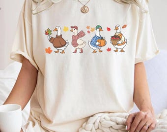 Puede incluir: Camiseta color crema con una ilustración de cuatro gansos vestidos con atuendos otoñales. Cada ganso sostiene objetos temáticos de otoño como hojas y calabazas. Los gansos están en fila, con hojas coloridas esparcidas.