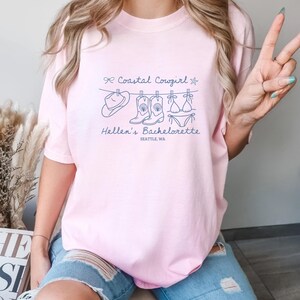 Peut inclure: T-shirt rose clair avec un dessin au trait bleu d'un chapeau, de bottes et d'un bikini suspendus à une corde à linge. Le texte indique "Coastal Cowgirl" et "Hellen's Bachelorette, Seattle, WA."