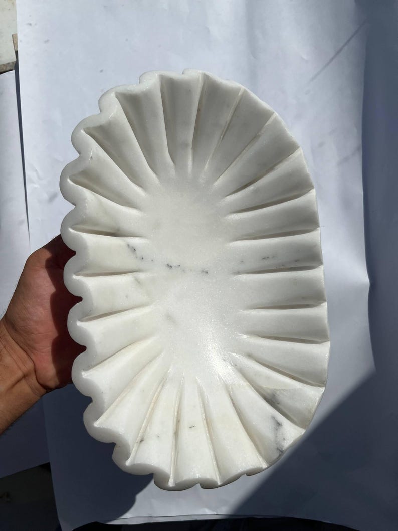 May include: A white marble decorative bowl with a scalloped edge. The bowl has a fan-like design with carved lines radiating from the center. The bowl is held by a hand and is set against a white background.