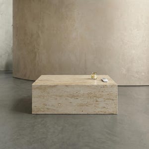 Può includere: Un tavolino rettangolare in pietra beige con una candela e un telecomando sopra. Il tavolo è un blocco solido di pietra, con una superficie strutturata. Lo sfondo è un muro marrone chiaro neutro.