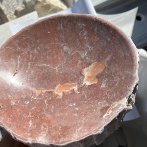 Puede incluir: Un cuenco de piedra de color marr&oacute;n rojizo, de forma irregular, con un interior poco profundo y redondeado. El cuenco tiene una superficie rugosa y texturizada con parches de color m&aacute;s claro y un borde ligeramente elevado. El cuenco parece estar tallado a mano.