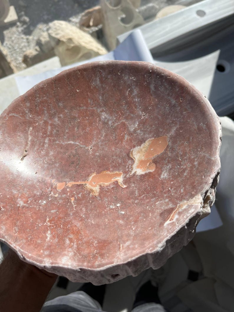 Puede incluir: Un cuenco de piedra de color marr&oacute;n rojizo, de forma irregular, con un interior poco profundo y redondeado. El cuenco tiene una superficie rugosa y texturizada con parches de color m&aacute;s claro y un borde ligeramente elevado. El cuenco parece estar tallado a mano.