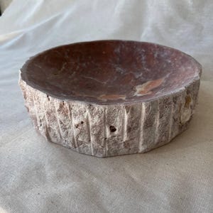 Puede incluir: Un cuenco redondo hecho a mano con un exterior rugoso y texturizado y un interior liso de color marr&oacute;n rojizo. El borde del cuenco tiene un aspecto natural e inacabado, mostrando la materia prima. El cuenco es probablemente de piedra o material similar.