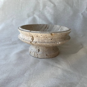 Può includere: Una ciotola a piedistallo in pietra beige con un bacino ampio e poco profondo. La ciotola ha una superficie strutturata con piccoli fori e un design decorativo a più livelli attorno alla base. È un oggetto decorativo.