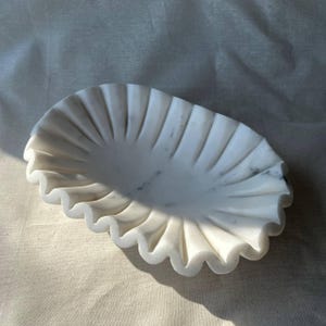 May include: A white marble decorative dish with a scalloped edge. The dish has a wavy, ruffled design and is made of white marble with subtle gray veining. The dish is oval-shaped and sits on a neutral-colored surface.
