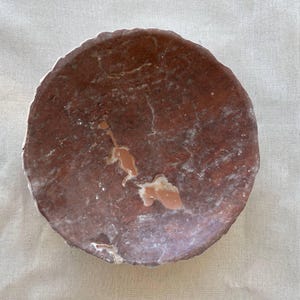 Puede incluir: Un plato redondo de piedra de color marr&oacute;n rojizo con una superficie rugosa y texturizada. El plato tiene un aspecto natural y org&aacute;nico con vetas y parches de color m&aacute;s claro. Los bordes son irregulares, lo que a&ntilde;ade encanto r&uacute;stico.