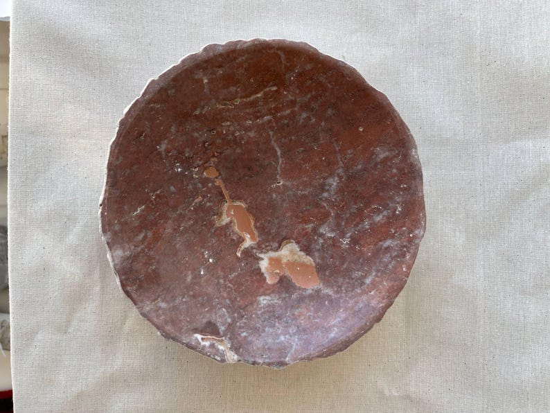 Puede incluir: Un plato redondo de piedra de color marr&oacute;n rojizo con una superficie rugosa y texturizada. El plato tiene un aspecto natural y org&aacute;nico con vetas y parches de color m&aacute;s claro. Los bordes son irregulares, lo que a&ntilde;ade encanto r&uacute;stico.