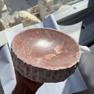 Puede incluir: Un cuenco de piedra redondo tallado a mano, de tonos rojizos. El cuenco tiene un exterior rugoso y texturizado y un interior liso con vetas de color m&aacute;s claro. El cuenco tiene aproximadamente 20 cm de di&aacute;metro y probablemente se utiliza con fines decorativos.