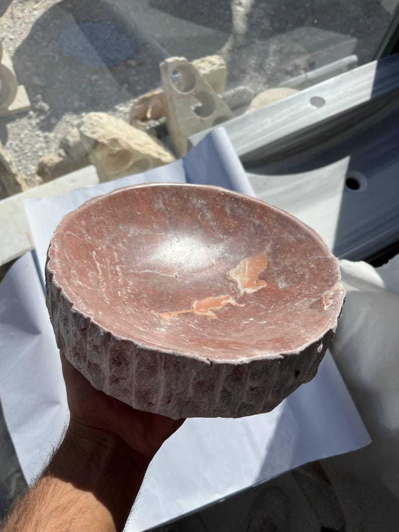 Puede incluir: Un cuenco de piedra redondo tallado a mano, de tonos rojizos. El cuenco tiene un exterior rugoso y texturizado y un interior liso con vetas de color m&aacute;s claro. El cuenco tiene aproximadamente 20 cm de di&aacute;metro y probablemente se utiliza con fines decorativos.