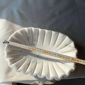 May include: A white marble oval dish with a scalloped edge. A ruler shows the dish is approximately 12 inches long. The dish has a fluted design and is set on a white surface.