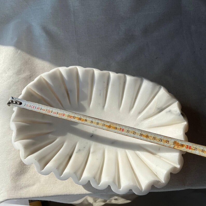 May include: A white marble oval dish with a scalloped edge. A ruler shows the dish is approximately 12 inches long. The dish has a fluted design and is set on a white surface.